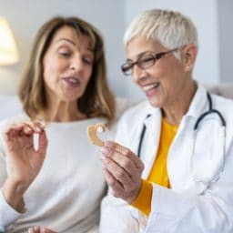 Mature female doctor hearing specialist in her office trying hearing aid equipment to a patient elderly senior woman. The doctor laryngologist explains to senior woman how to wear a hearing aid